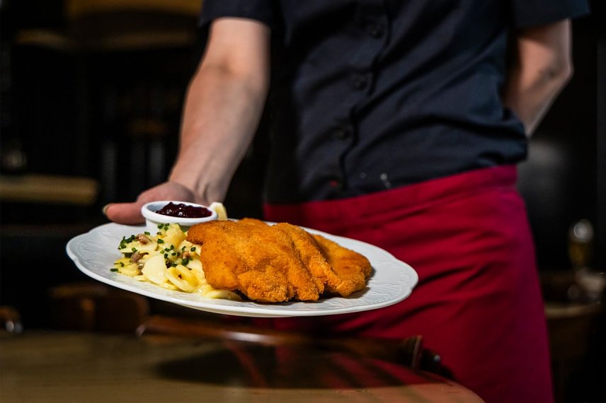 Bild: Eine Kellnerin mit roter Sch&uuml;rze h&auml;lt einen Teller mit Schnitzel und Kartoffeln in die Kamera. 