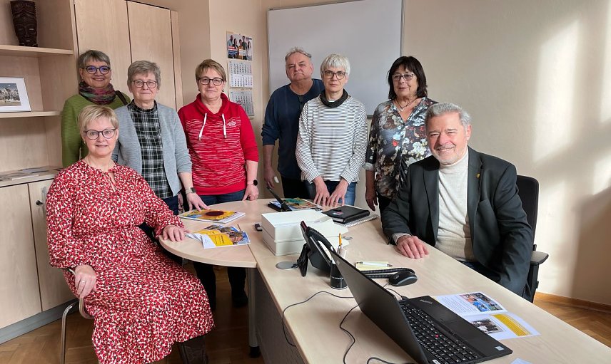 Bild: Mehrere Menschen sitzen und stehen an einem Schreibstisch in einem B&uuml;ro. 